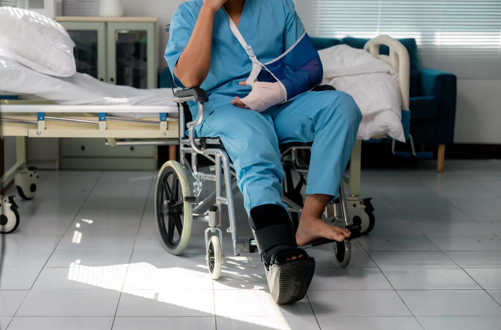 Injured patient sitting in a wheelchair beside a hospital bed, wearing an arm sling and ankle brace while recovering from surgery or an accident, focused on rehabilitation